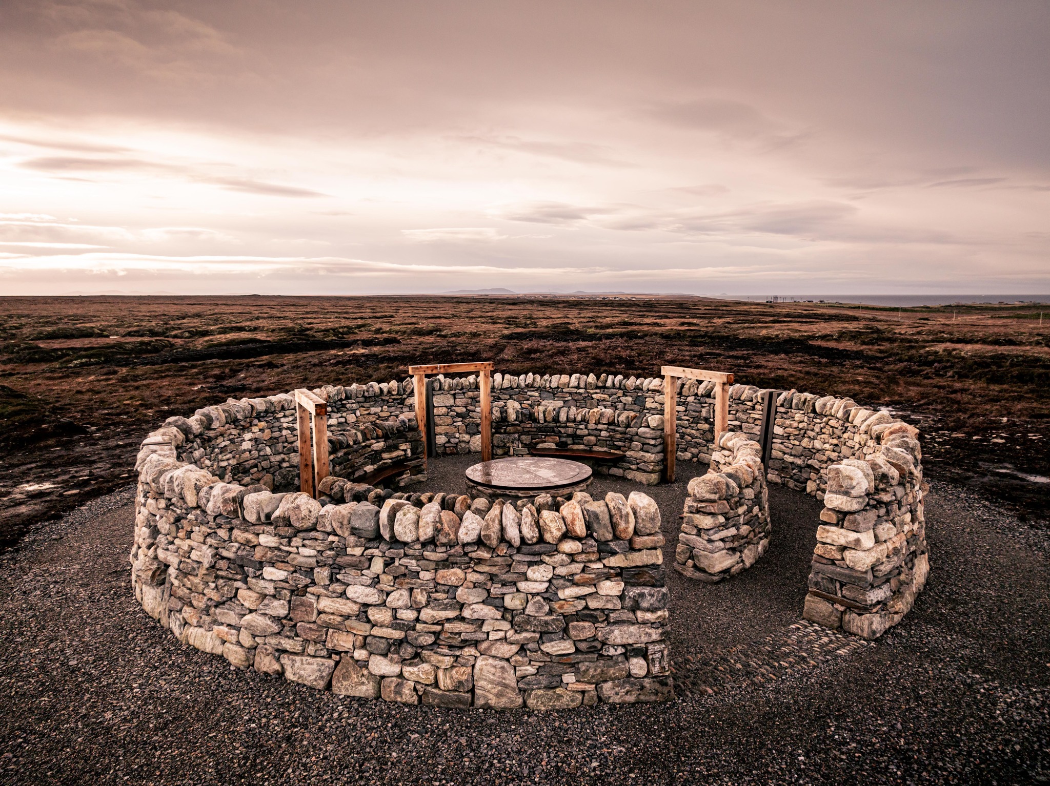 An aerial view of a circular stone structure, named Na Dorsan, made of concentric rings of stacked rocks. Inside the rings are curved wooden benches spaced evenly around a central metal disk set into the ground. The entire arrangement sits on gravel, with darker earth surrounding the circular layout.