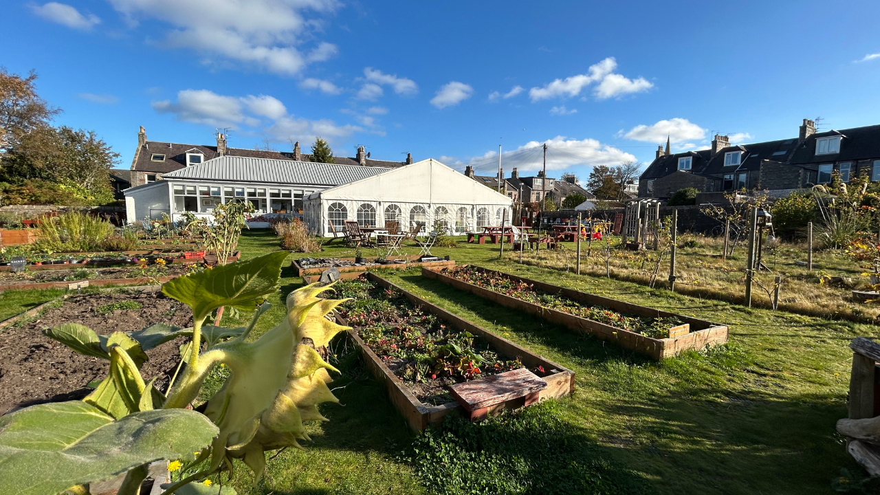 Raised garden beds with plants in front of a white greenhouse, under a blue sky; "The Gordon &amp; Ena Baxter Foundation" logo is overlaid at the top.