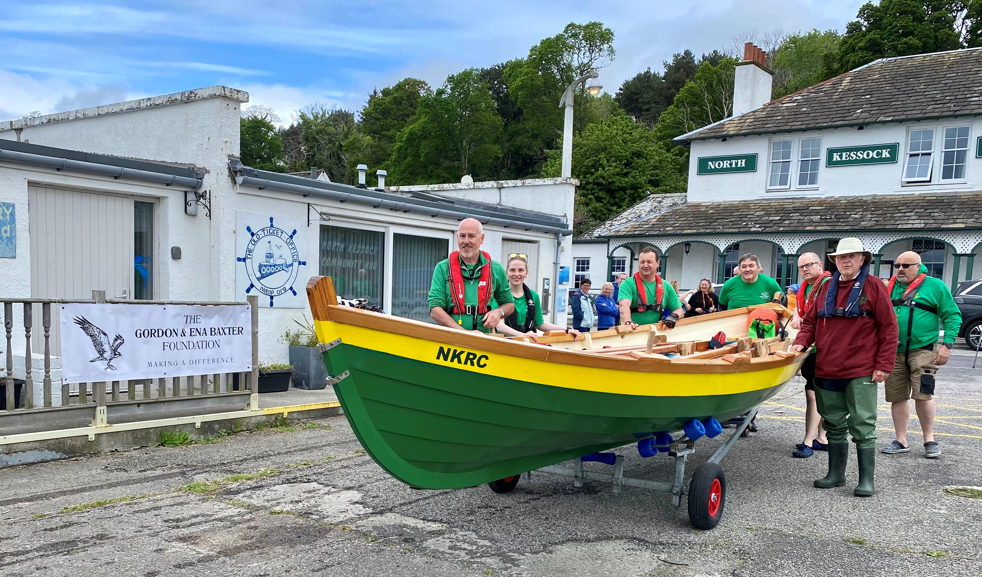 A crew of men and women stand next to a coastal rowing skiff, which is upon a road trailer. A green sign on a building in the background states North Kessock. Next to the boat is a banner for The Gordon and Ena Baxter Foundation. 