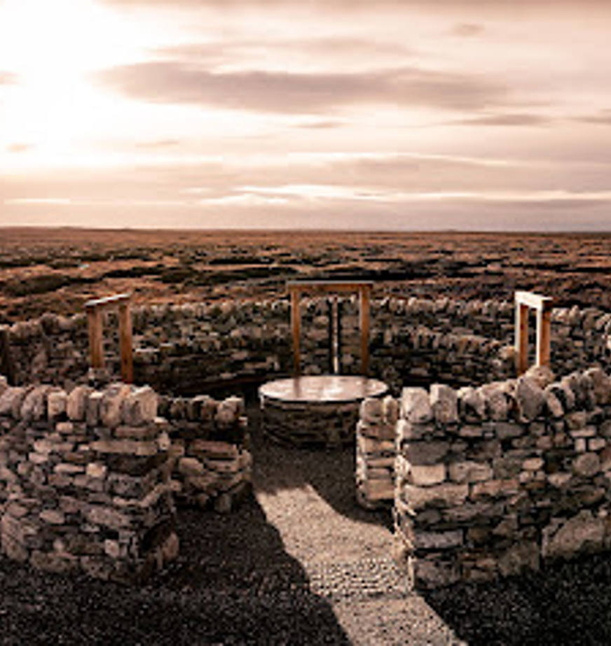 An aerial view of a circular stone structure, named Na Dorsan, made of concentric rings of stacked rocks. Inside the rings are curved wooden benches spaced evenly around a central metal disk set into the ground. The entire arrangement sits on gravel, with darker earth surrounding the circular layout.