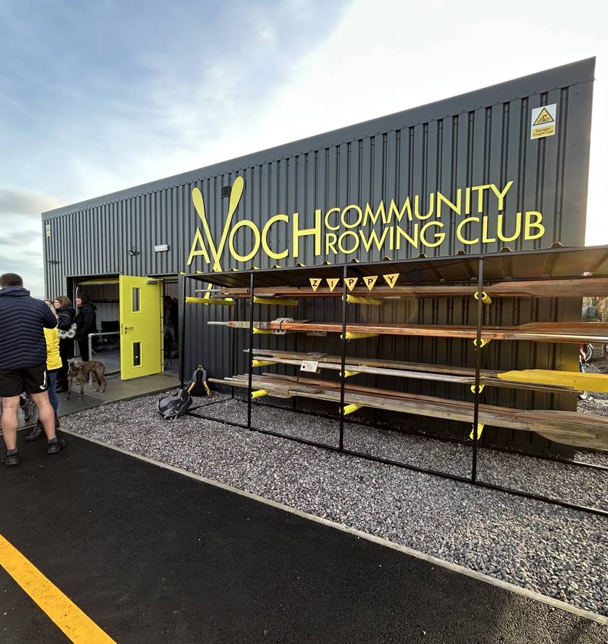 The entrance to Avoch Community Rowing Club, featuring a modern building with large yellow lettering. Several people are gathered outside. A rack of rowing oars is visible to the side, and the scene is illuminated by soft lighting under a partly cloudy sky.