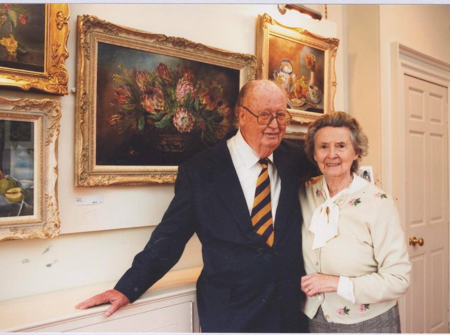 An older couple smile at the camera, pictured in front of some still life paintings of flowers which are hung in a brightly lit room. 